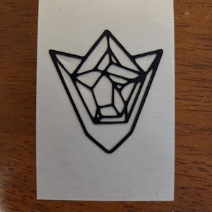 May include: A temporary tattoo featuring a black, geometric, angular design that resembles a stylized crystal or gemstone shape. The graphic is printed on a rectangular white backing, showcasing sharp, clean lines and a symmetrical, multifaceted appearance.