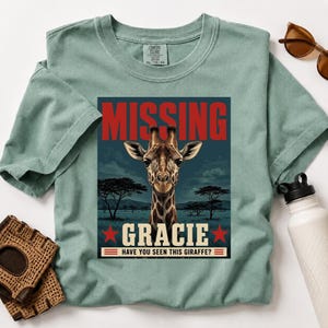 May include: A sage green t-shirt featuring a graphic print of a giraffe in a savanna landscape. The text reads MISSING GRACIE HAVE YOU SEEN THIS GIRAFFE? The design includes red stars and horizontal lines, set against a dark blue rectangular background.