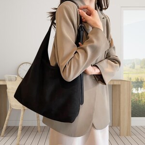 May include: A large, black, soft-textured hobo-style shoulder bag with a single wide strap and a silver-toned metal buckle adjustment feature. The bag has a slouchy, unstructured silhouette and a smooth, matte finish, hanging from a shoulder.