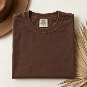 May include: A solid dark brown short-sleeve t-shirt with a crew neckline. The inner neck label displays the text Comfort Colors and the size L G G. The shirt is folded neatly, showcasing a smooth, matte fabric texture and a consistent deep brown color.