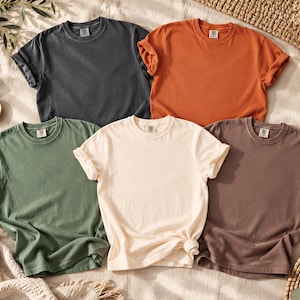 May include: A collection of five short-sleeved crew neck t-shirts in solid colors including charcoal gray, burnt orange, sage green, cream, and mocha brown. Each shirt features a standard collar and rolled sleeves, arranged in a group.