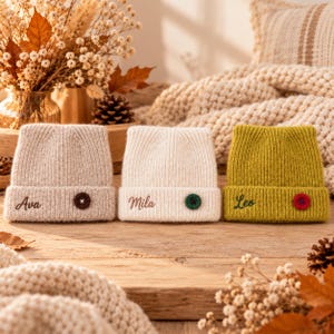 Peut inclure: Trois bonnets en tricot côtelé de couleur beige, crème et vert olive. Chaque bonnet présente un revers avec un prénom brodé et un bouton décoratif. Les prénoms Ava, Mila et Leo sont cousus en fil foncé sur chaque bonnet respectif.