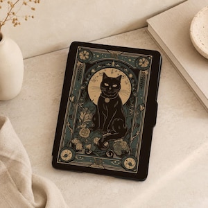 May include: A black e-reader case featuring a central illustration of a black cat sitting within a circular frame, surrounded by floral motifs and intricate geometric borders in shades of muted blue, cream, and dark brown on a black background.