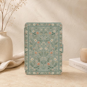 Puede incluir: Una funda rectangular para lector electrónico con fondo verde salvia y un patrón botánico repetitivo de pequeñas flores silvestres blancas, amarillas y rosas con follaje verde. La funda tiene un borde fino y una pequeña pestaña lateral.