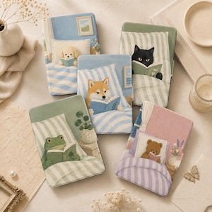 Puede incluir: Cinco fundas rectangulares para lectores electrónicos con ilustraciones de animales leyendo en la cama. Los diseños incluyen un cachorro crema, un gato negro, un Shiba Inu, una rana verde y un oso de peluche marrón bajo ropa de cama a rayas.