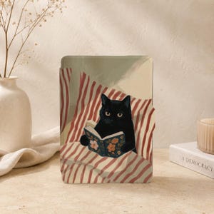 May include: A rectangular e-reader case featuring an illustration of a black cat sitting on a red and white striped surface while holding an open book with a blue cover decorated with orange and pink floral patterns.