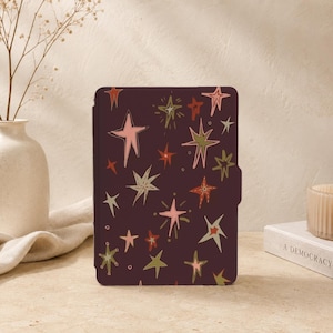 May include: A dark purple e-reader case featuring a repeating pattern of hand-drawn style stars in shades of light pink, olive green, and burnt orange. The stars vary in size and shape, scattered across the surface of the rectangular cover.