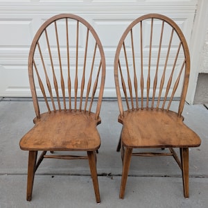 May include: Two wooden Windsor-style chairs with a natural finish. Each chair features a curved back with vertical spindles and a contoured seat. The chairs are made of wood and have a classic design.