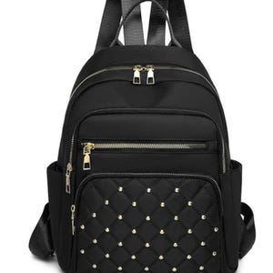 May include: A black backpack featuring a quilted front pocket adorned with small silver-toned studs. The bag includes a main zippered compartment, a secondary front zippered pocket, two side slip pockets, and adjustable black shoulder straps.