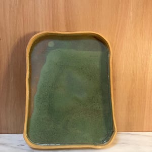 May include: A rectangular ceramic tray with rounded corners and a raised edge. The interior surface features a mottled green glaze with a smooth, glossy finish, while the outer rim displays a natural, unglazed light tan clay texture.
