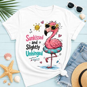 May include: A white short-sleeve t-shirt featuring a graphic of a pink flamingo wearing black sunglasses and a flower, standing in a blue patterned inner tube. The text reads Sunkissed and Slightly Unhinged, accompanied by a yellow sun and hearts.