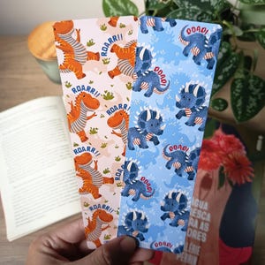 Pode incluir: Dois marcadores de página retangulares com ilustrações de dinossauros. O da esquerda é rosa claro com dinossauros laranja e o texto ROARR!!. O da direita é azul claro com triceratops azul escuro e o texto ROAR!