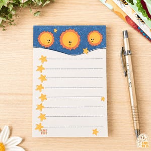 Pode incluir: Um bloco de notas retangular com um cabeçalho azul apresentando três sóis amarelos com rostos e pequenas estrelas amarelas. A área de escrita branca tem linhas tracejadas horizontais e uma coluna vertical de estrelas amarelas. Texto Lari Reis.