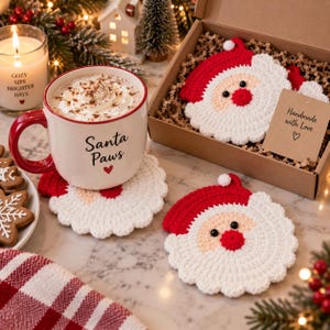 以下が含まれることがあります： 赤い持ち手とSanta Pawsの文字が入った白いセラミック製マグカップ。赤、白、ピーチ色のサンタクロースの形をした丸いかぎ針編みのコースターが3枚添えられています。1枚はマグカップの下に敷かれ、2枚はタグ付きのギフトボックスに入っています。