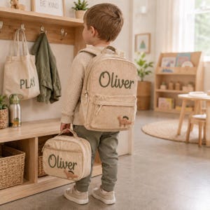 Può includere: Uno zaino e una borsa per il pranzo abbinata in velluto a coste beige, personalizzati con il nome Oliver ricamato in verde scuro e una piccola toppa a forma di dinosauro. Lo zaino presenta uno scomparto principale e una tasca frontale con cerniere dorate.