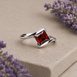 Könnte beinhalten: Ein silberfarbener Ring mit einem einzelnen tiefroten Edelstein im Prinzessschliff, der in eine Ringschiene im Bypass-Stil gefasst ist. Das Metallband schwingt sich um den quadratischen Stein und sorgt für ein glattes, poliertes Erscheinungsbild.