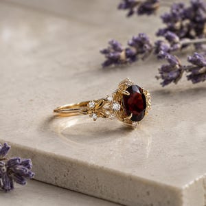 Op de afbeelding: Een gouden ring met een centrale, ovaal geslepen, dieprode granaat. De ringband is ontworpen met gedetailleerde gouden bladmotieven en kleine, heldere, rond geslepen accentdiamanten rondom de granaat, wat zorgt voor een verfijnde botanische zetting.