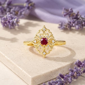 May include: A gold ring featuring a central round red gemstone surrounded by an intricate filigree metal frame. Small clear stones are embedded within the decorative metalwork, which has a pointed diamond shape. The band has a textured finish.