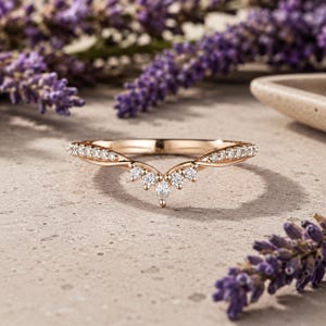 May include: A rose gold V-shaped ring featuring a series of small, round, clear stones set along the band, culminating in a central marquise-cut stone at the point of the V. The metal has a polished finish and a delicate, curved silhouette.