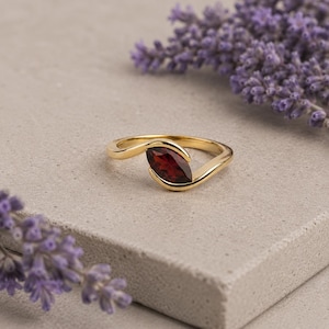 Könnte beinhalten: Ein Goldring mit einem einzelnen, tiefroten Granat-Edelstein im Marquise-Schliff, der in eine glatte Ringschiene im Bypass-Stil gefasst ist. Das Metall hat eine polierte Oberfläche und der Stein ist mittig zwischen den geschwungenen Enden platziert.