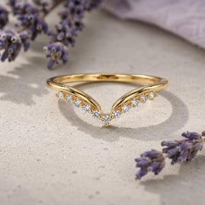 May include: A gold V-shaped chevron ring featuring a row of small, round, clear faceted stones set along the front curve. The band has a polished finish and a pointed center design, creating a delicate and symmetrical appearance for the jewelry piece.