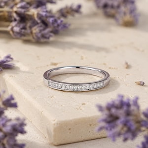 May include: A silver-toned metal band ring featuring a channel setting with a row of small, clear, round-cut stones embedded along the front half of the band. The ring has a polished, reflective surface and a slim, uniform width throughout its design.
