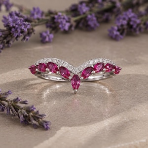 May include: A V-shaped ring crafted from polished silver-toned metal, featuring a row of marquise-cut vibrant pink gemstones set along the band, accented by a delicate line of small, clear round-cut stones embedded along the upper curved edge.