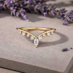 May include: A gold V-shaped ring featuring a central marquise-cut diamond flanked by six smaller round-cut diamonds set along the band. The band has a polished finish and follows a chevron silhouette, creating a pointed design with clear stones.