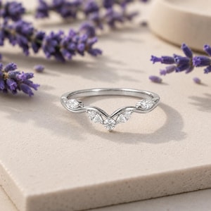 May include: A silver-toned ring featuring a curved band with a central round-cut stone flanked by two marquise-cut stones on each side, creating a symmetrical V-shaped design. The metal band has a polished finish and holds five clear, faceted stones.