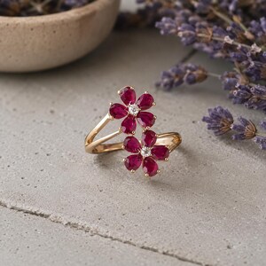 May include: A gold bypass ring featuring two flower-shaped motifs. Each flower is composed of five teardrop-shaped deep red rubies surrounding a single small, clear round-cut diamond at the centre. The band is polished gold with a smooth finish.