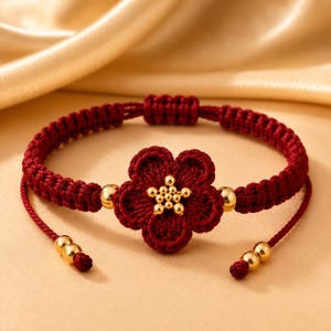 May include: A deep red woven cord bracelet featuring a central five-petaled flower motif made of matching red thread. The center of the flower is adorned with a cluster of small gold-toned beads, with additional gold beads accenting the cord ends.
