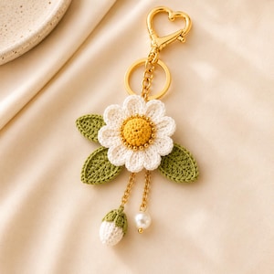 May include: A crochet daisy keychain featuring a white flower with a yellow center and green leaves. The accessory includes a gold-toned heart-shaped clasp, a circular ring, and two dangling gold chains, one ending in a small white bud and one in a pearl.