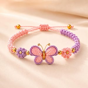 May include: A woven cord bracelet featuring a central butterfly charm with pink and purple thread, flanked by two small flower-shaped beads. The band consists of alternating pink and purple braided cord sections accented with small gold-toned metal beads.