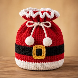 May include: A red crochet drawstring bag featuring a white ruffled top edge, a black horizontal band across the centre, and a yellow rectangular buckle detail. The bag includes a cream-coloured drawstring cord with two small pom-poms at the ends.