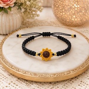 May include: A black macrame cord bracelet featuring a central crocheted sunflower charm with yellow petals and a brown center. The bracelet includes two small gold-toned metallic beads flanking the sunflower and an adjustable sliding knot closure.