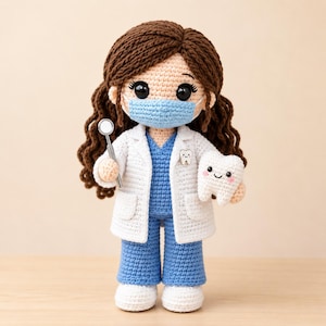 May include: A crocheted doll dressed as a dentist with long brown hair, wearing a white lab coat over blue scrubs and a light blue face mask. The doll holds a small silver dental mirror in one hand and a white plush tooth with a face in the other.