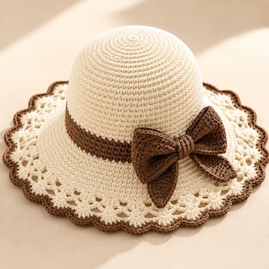 May include: A cream-colored crocheted sun hat featuring a dark brown band around the crown and a large, matching dark brown crocheted bow on the side. The wide brim has an intricate, open-work lace pattern finished with a dark brown scalloped edge.