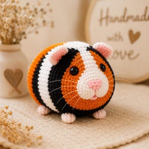 Può includere: Un peluche a forma di porcellino d'India realizzato all'uncinetto, di forma rotonda, con un motivo tricolore in filato arancione, nero e bianco. Il pupazzo ha piccole orecchie rosa, naso rosa, occhi a perla neri e sottili baffi bianchi.