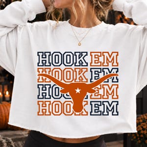 Könnte beinhalten: Ein weißes Langarm-Sweatshirt mit einem Grafikdruck, der den Text HOOK EM in einem Rastermuster aus marineblauen und rostorangen Blockbuchstaben wiederholt. In der Mitte befindet sich eine große, rostorange Longhorn-Silhouette mit einem weißen Stern.