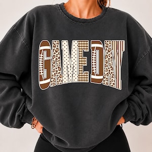 May include: A charcoal grey crewneck sweatshirt featuring the text GAME DAY in a large, arched font. The letters incorporate various patterns including brown football shapes, tan leopard print, white polka dots, brown plaid, and vertical stripes.