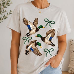 May include: A light gray short-sleeve t-shirt featuring a graphic print of three mallard ducks in flight with green heads, brown and white wings, and blue wing patches, interspersed with three dark green hand-drawn style ribbon bows.