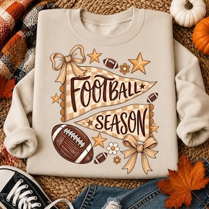 Könnte beinhalten: Ein beigefarbenes Sweatshirt mit Rundhalsausschnitt und einem Grafikdruck von zwei karierten Wimpeln mit dem Text "FOOTBALL SEASON" in brauner Schrift. Das Design umfasst braune Footballs, beigefarbene Sterne, kleine weiße Blumen und zwei Schleifen.