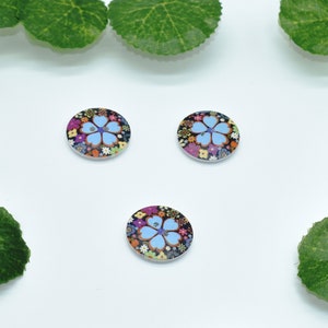 May include: Three round wooden buttons with a floral design. The buttons feature a blue flower with a pink center surrounded by colorful flowers on a black background.