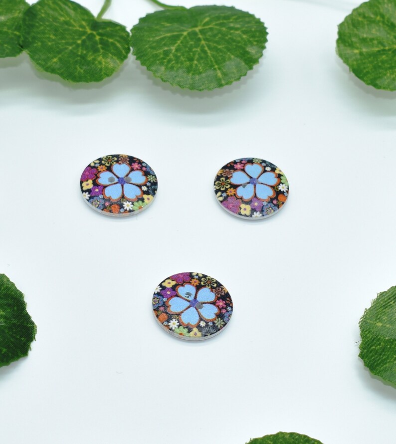 May include: Three round wooden buttons with a floral design. The buttons feature a blue flower with a pink center surrounded by colorful flowers on a black background.