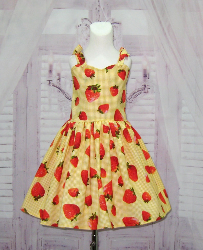 May include: A yellow and white gingham dress with a strawberry print. The dress has thin straps and a full skirt.