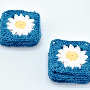 May include: Two stacks of square crochet coasters. Each coaster features a white daisy with a yellow centre, set against a teal blue background. The coasters are handmade and have a textured appearance.