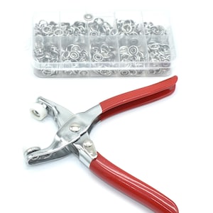 Puede incluir: Un juego de ojales de metal plateado y una herramienta para colocar ojales con mango rojo. Los ojales están en un organizador de plástico transparente con múltiples compartimentos. La herramienta para colocar ojales es de metal plateado y tiene un mango rojo.