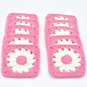 May include: Two stacks of pink and white crochet squares. Each square has a white flower design in the centre, framed by a pink border. The squares are arranged in neat rows, showing the handmade texture.
