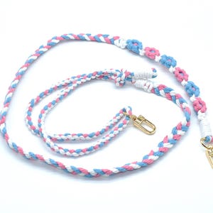 May include: A braided strap in white, pink, and light blue, with gold-colored clasps. The strap features a floral design in pink and light blue. The strap is designed for a bag or purse.