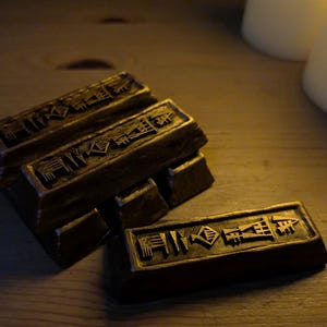 Puede incluir: Un conjunto de cuatro barras rectangulares de color bronce con bordes biselados. Cada barra presenta una serie de símbolos geométricos y angulares en relieve en la superficie superior, que recuerdan a una escritura antigua o marcas estilizadas.